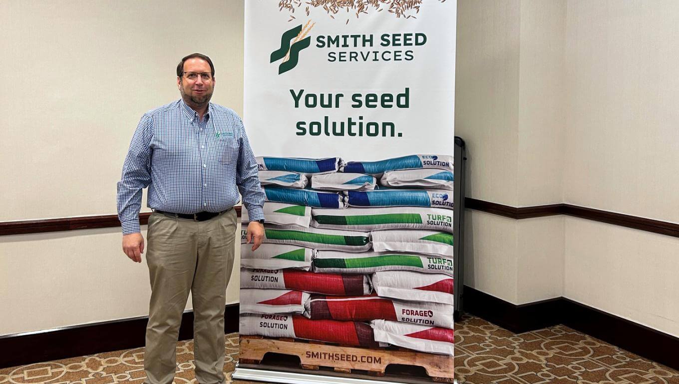 Man standing indoors next to a Smith Seed Services banner featuring the slogan 'Your seed solution' and stacked colorful seed bags, likely at a trade show or conference.