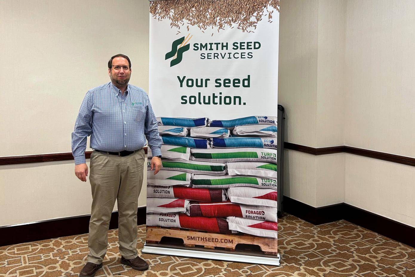 Man standing indoors next to a Smith Seed Services banner featuring the slogan 'Your seed solution' and stacked colorful seed bags, likely at a trade show or conference.