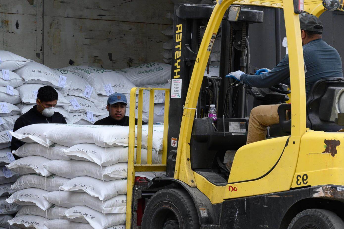 A yellow forklift lifts a stack of white bags while a worker in a hat and jacket operates it. Two other workers stand nearby among more stacked bags in a train boxcar.