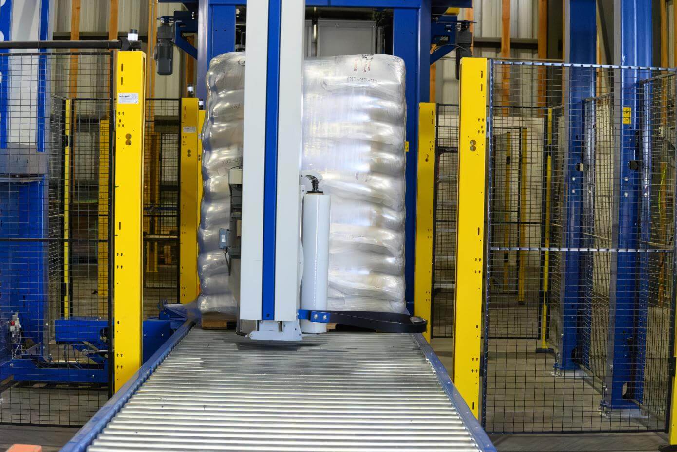 Automated pallet‑wrapping machine securing a tall stack of packaged goods with clear stretch film on a conveyor in an industrial warehouse.